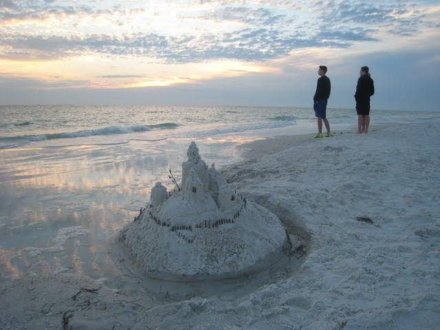 Two boys, standing beside a sandcastle built in white Florida sand, look out into the ocean.